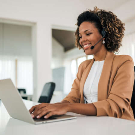 Smiling latin businesswoman working with laptop and headset, portrait. Cheerful assistant in the bright office.